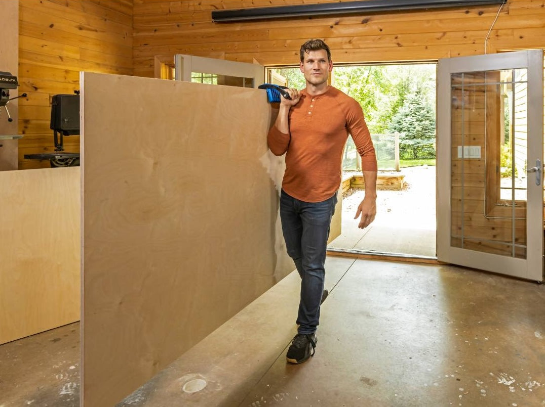 Man carrying large plywood panel with ergonomic blue and black clamp in woodworking tool accessories collection