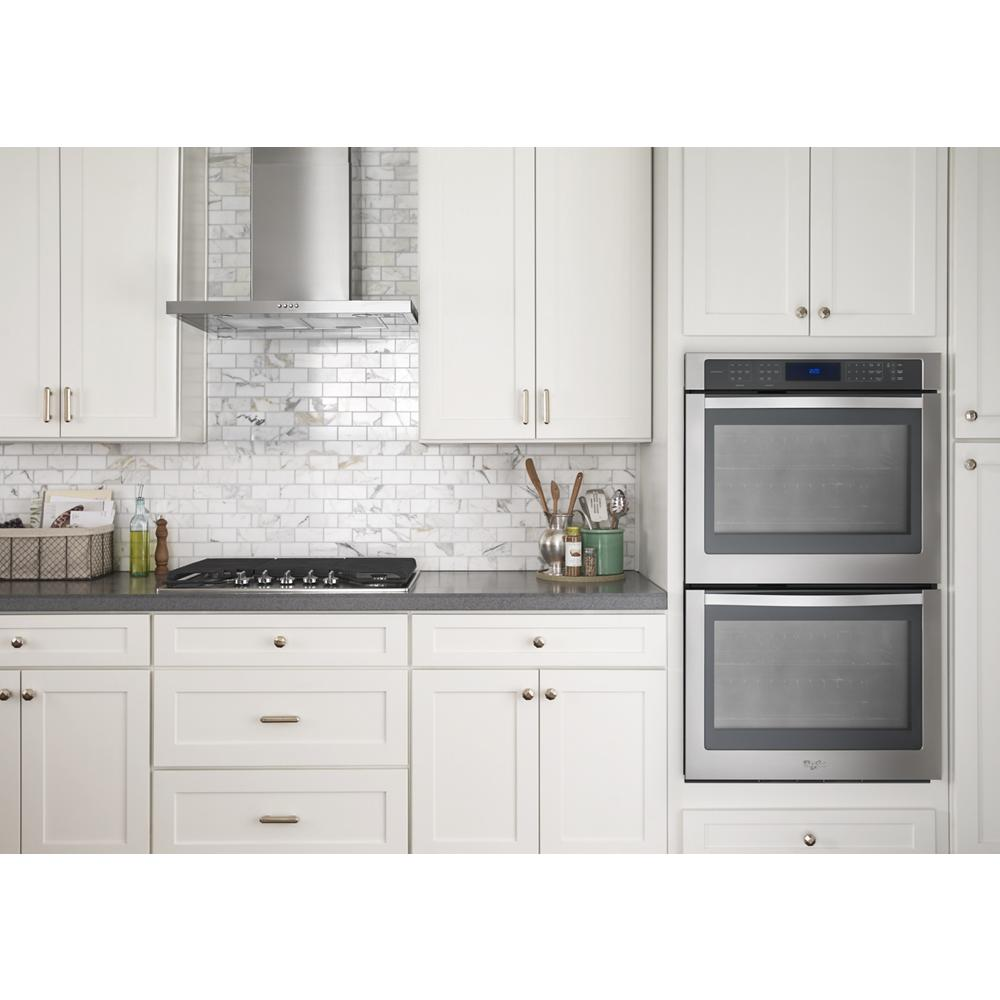 Modern kitchen scene featuring a stainless steel wall mount flat range hood above a gas cooktop with white cabinetry and double ovens.