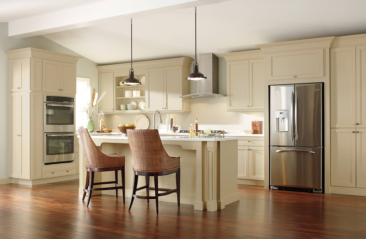 Bright kitchen with cream-colored cabinetry, stainless steel refrigerator and double oven, wooden flooring, and two wicker chairs at a central island.
