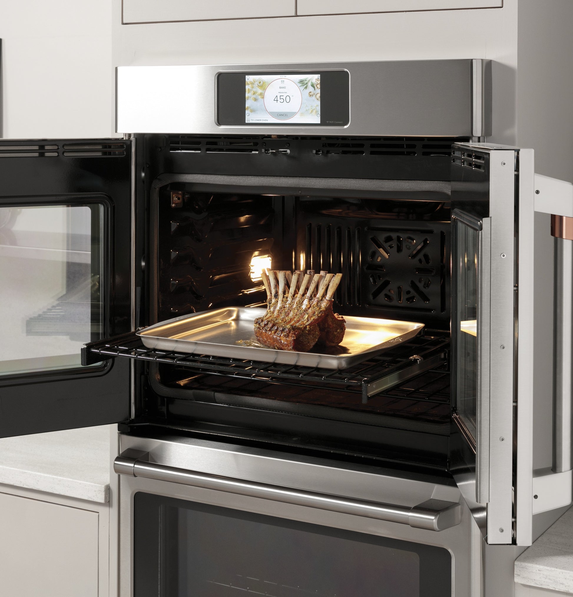 Open stainless steel double wall oven with digital display and rack holding a tray of roasting ribs in modern kitchen
