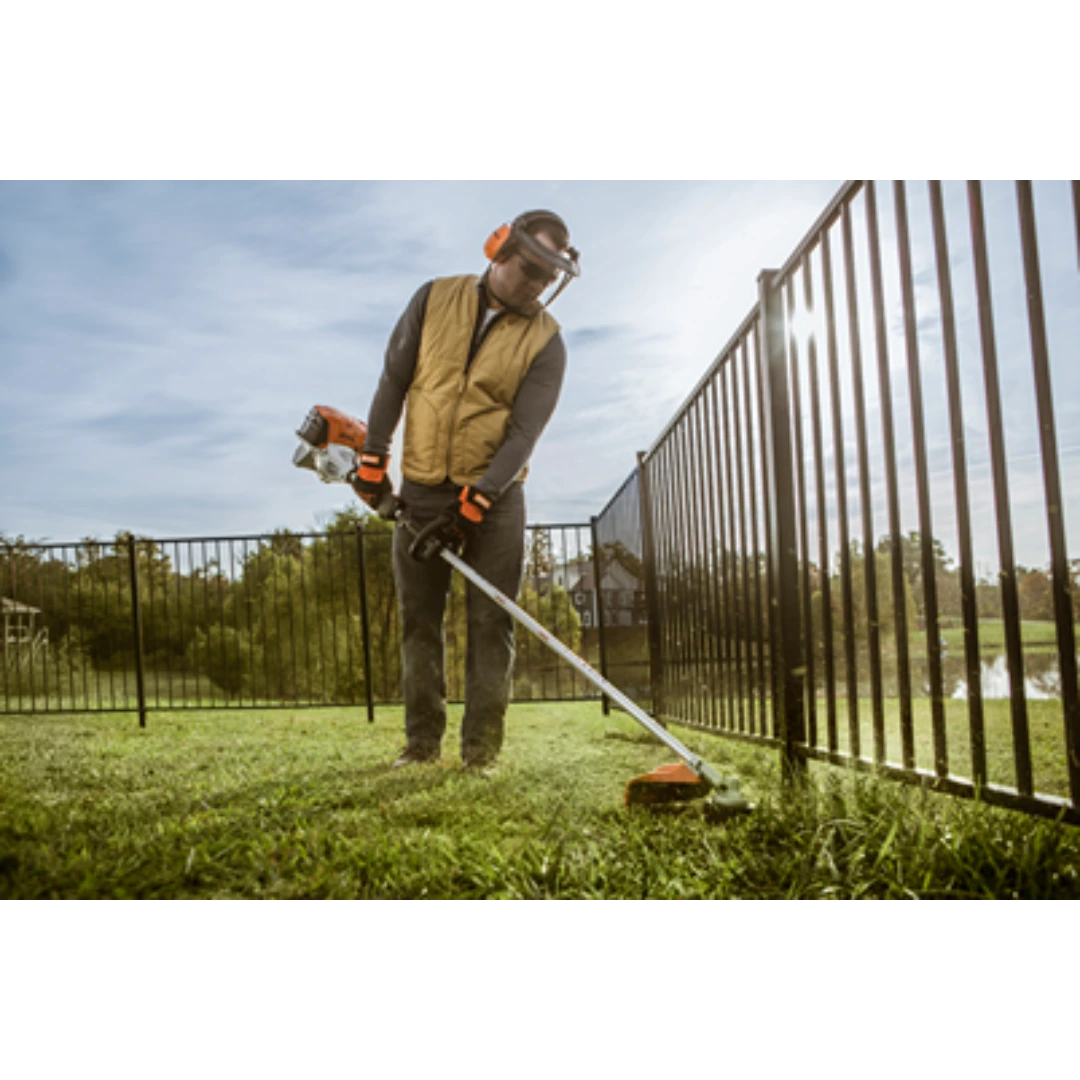 Person using a gas-powered loop handle trimmer with protective gear to edge grass near a metal fence on a sunny day