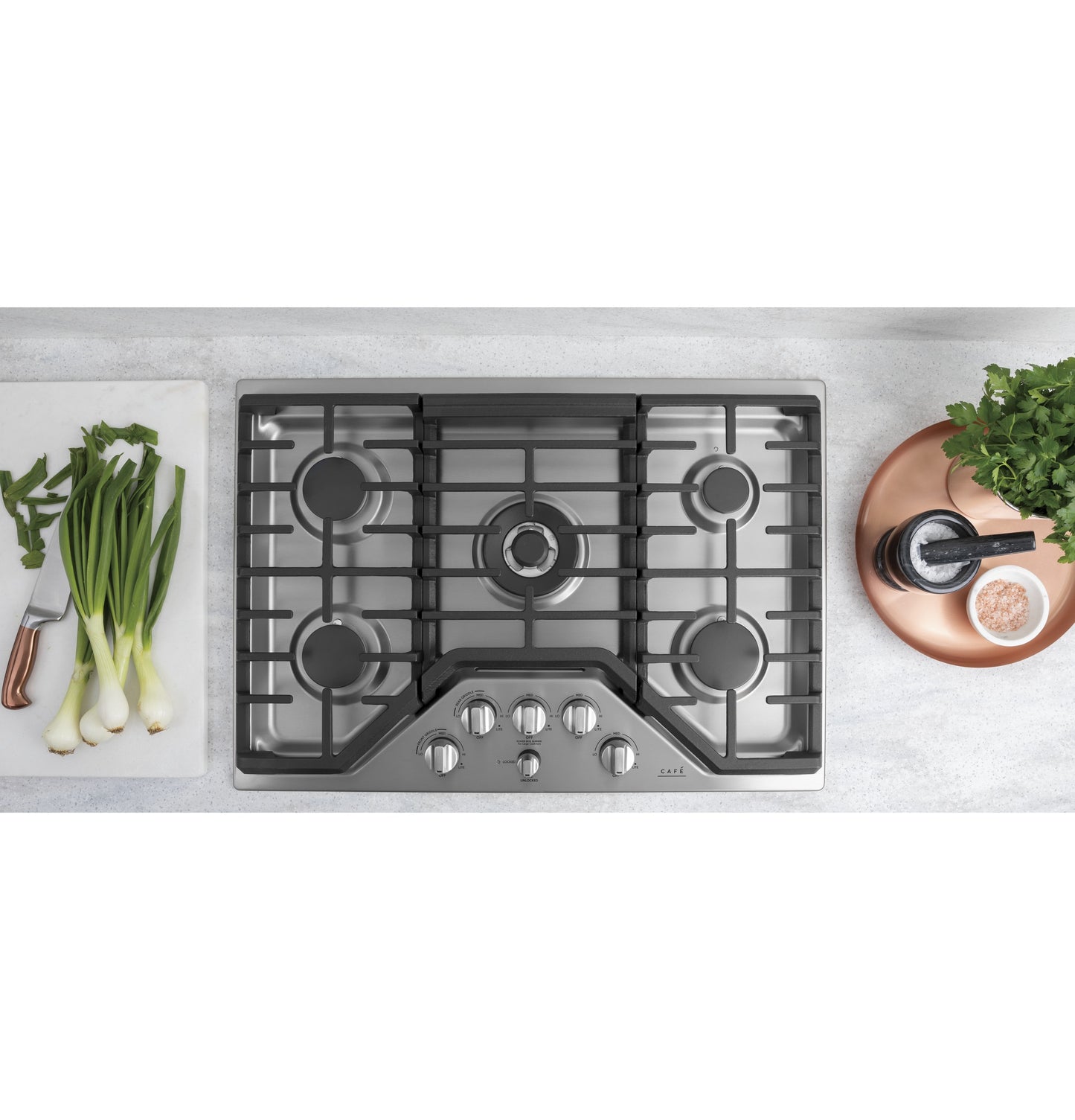 Stainless steel gas cooktop with five burners and metallic control knobs on a kitchen countertop with fresh herbs and spices nearby