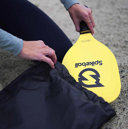 Yellow Spikeball paddle with black handle being placed into a black drawstring bag for outdoor games collection