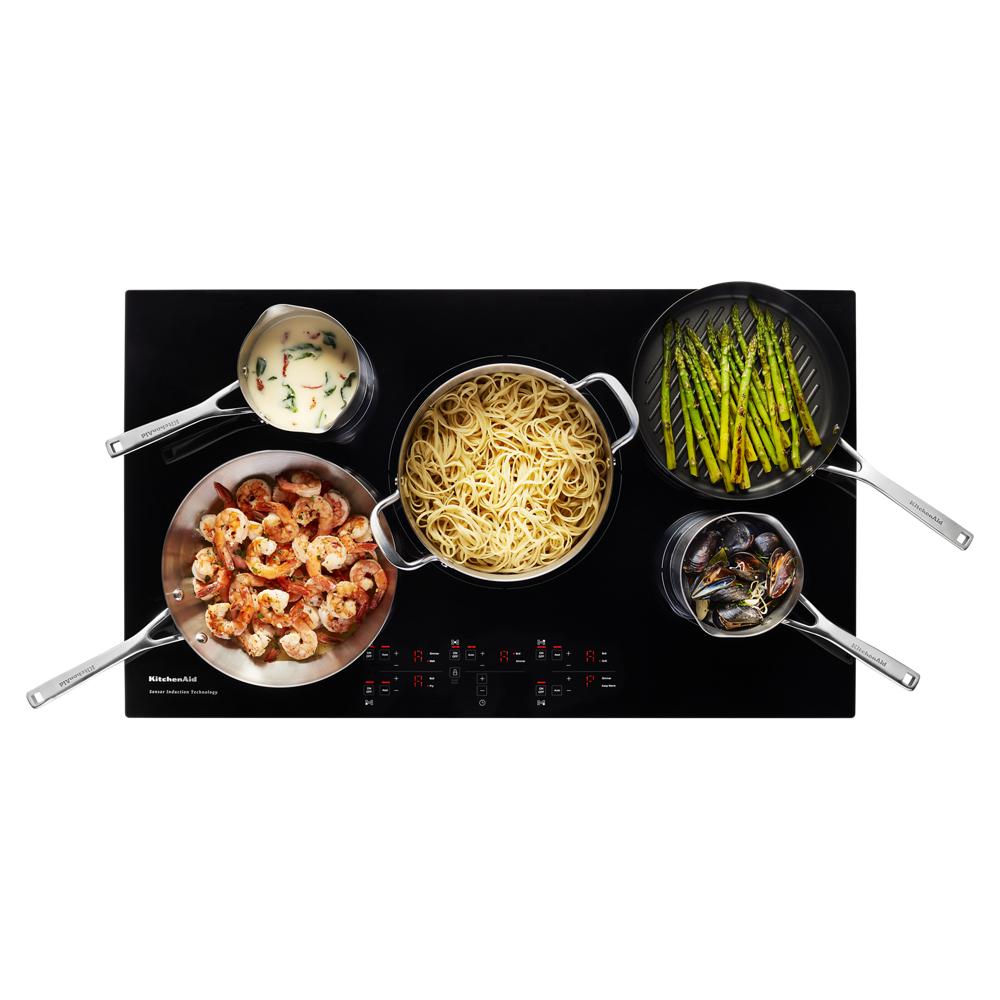Black 36-inch cooktop with five elements cooking shrimp, pasta, asparagus, sauce, and mussels in stainless cookware