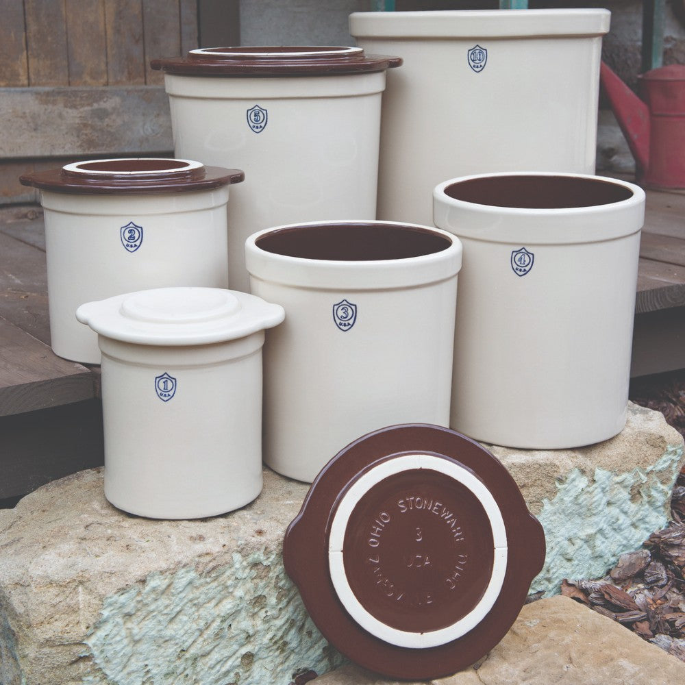 Collection of mixed-size beige stoneware crocks with dark brown interiors and small blue shield emblems outdoors on stone steps