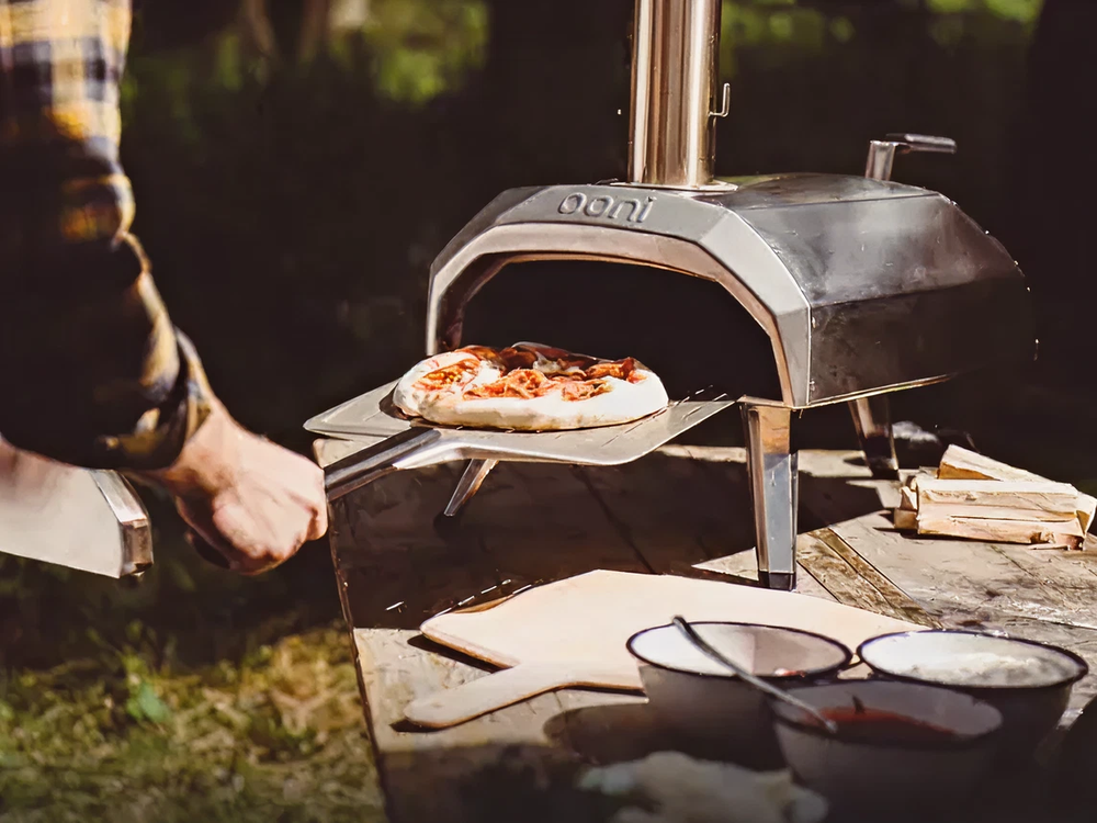 Person sliding a freshly baked pizza with toppings out of a stainless steel outdoor pizza oven set on a wooden table with prep bowls nearby.