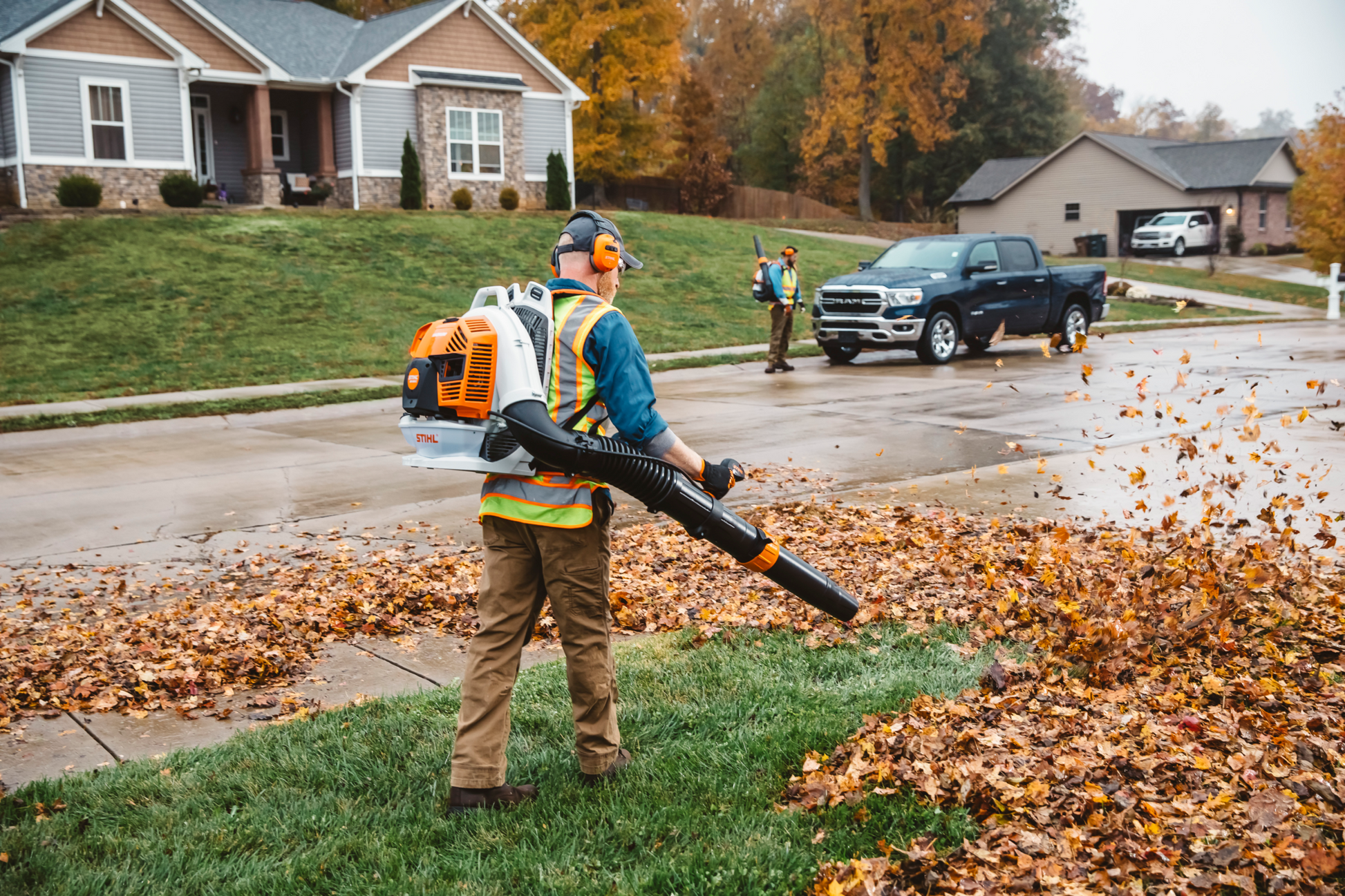 Worker using an orange and white backpack blower to clear leaves on a suburban lawn during autumn cleanup tasks