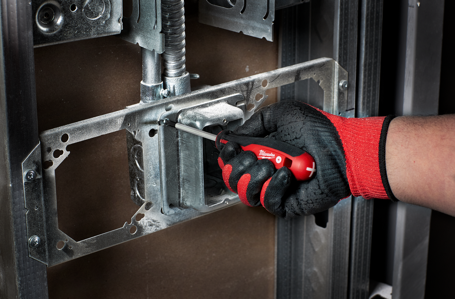 Hand wearing red and black glove using red screwdriver with ergonomic handle to install electrical box frame in home improvement setting
