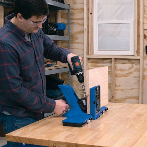 Man using cordless drill with blue pocket hole jig on hardwood bench top in woodworking tool accessories setting