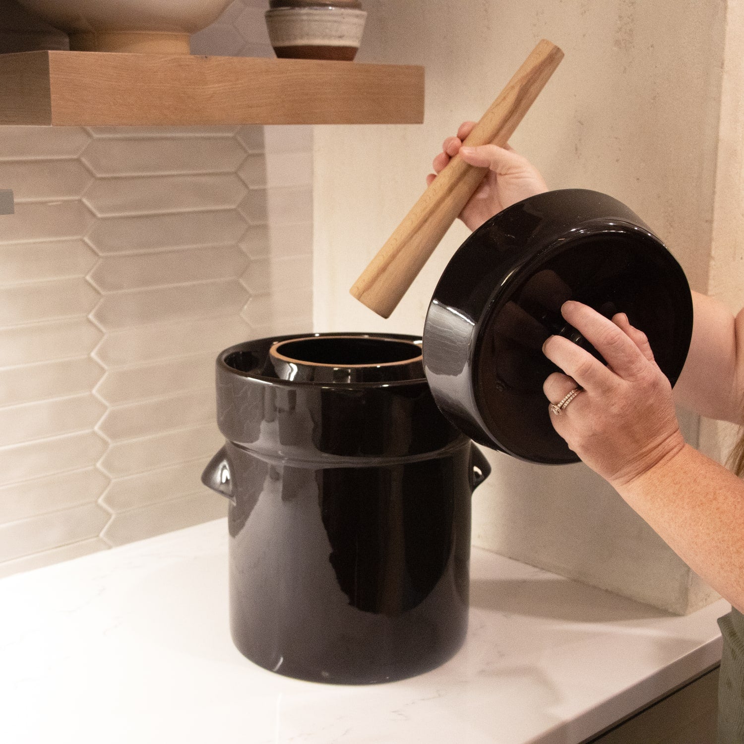 Hands placing wooden stomper into glossy black crock with lid on white countertop in kitchenware setting