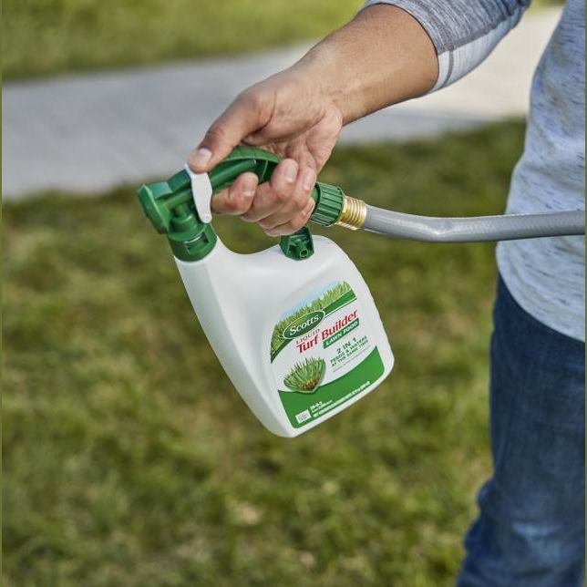 Person holding a green and white Scotts liquid lawn fertilizer sprayer attached to a garden hose in lawn care setting