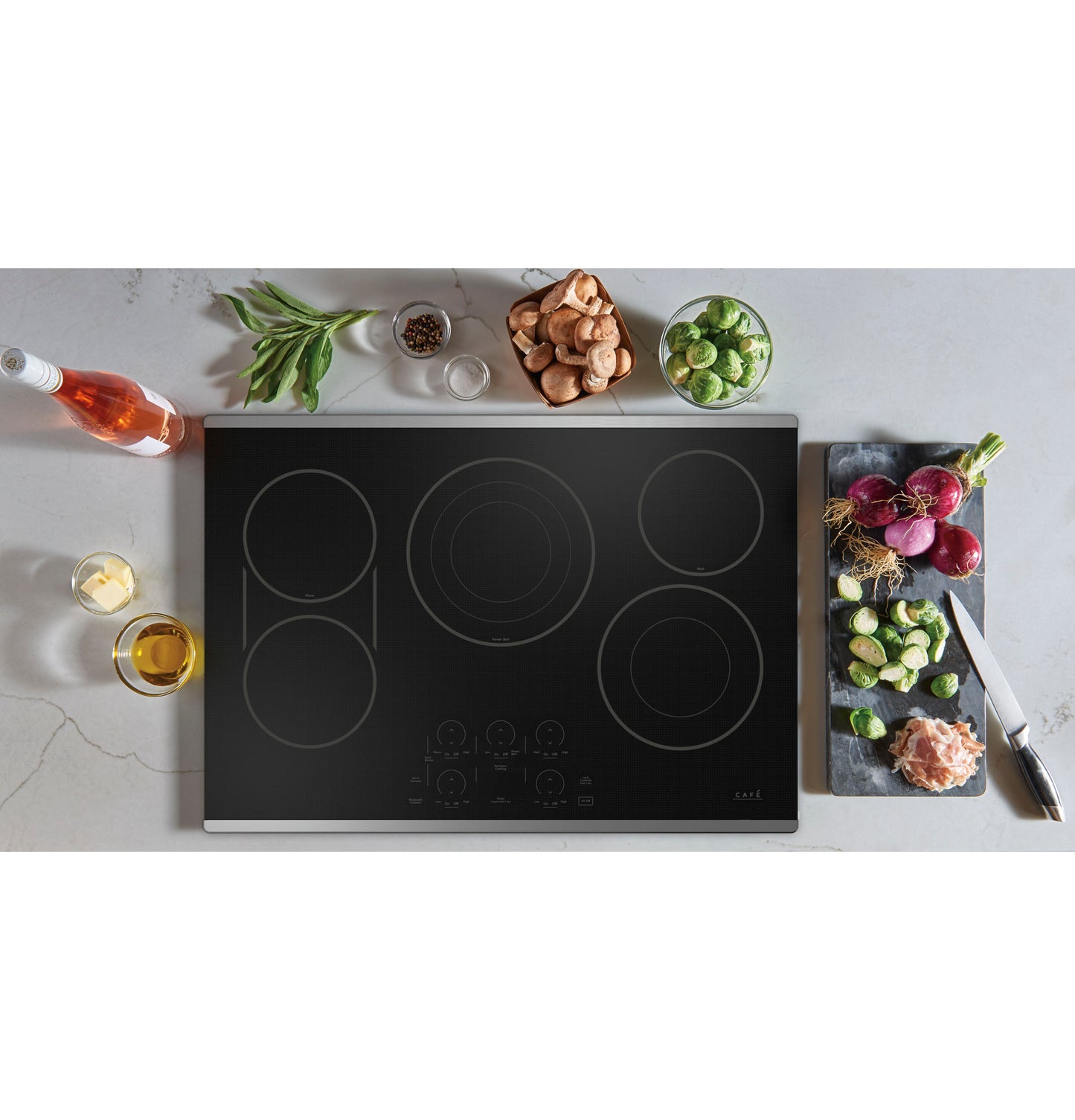Black glass electric cooktop with five burners and touch controls surrounded by fresh vegetables and cooking ingredients