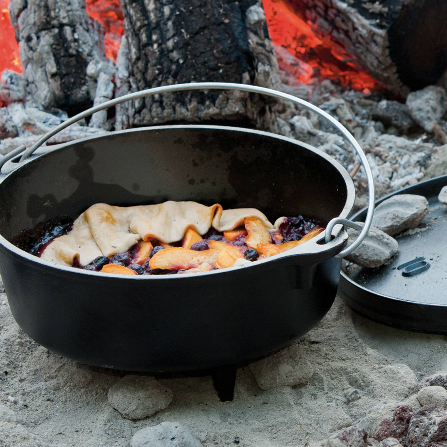 Pre-seasoned cast iron camp Dutch oven with lid set on campfire coals cooking fruit cobbler in kitchenware collection