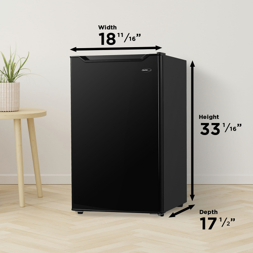 Compact black refrigerator with dimensions illustrated, standing next to a wooden side table and plant in kitchen appliance setting