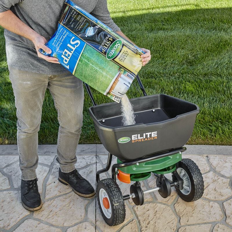 Person pouring granular lawn food into a Scotts Elite spreader on patio near green grass lawn