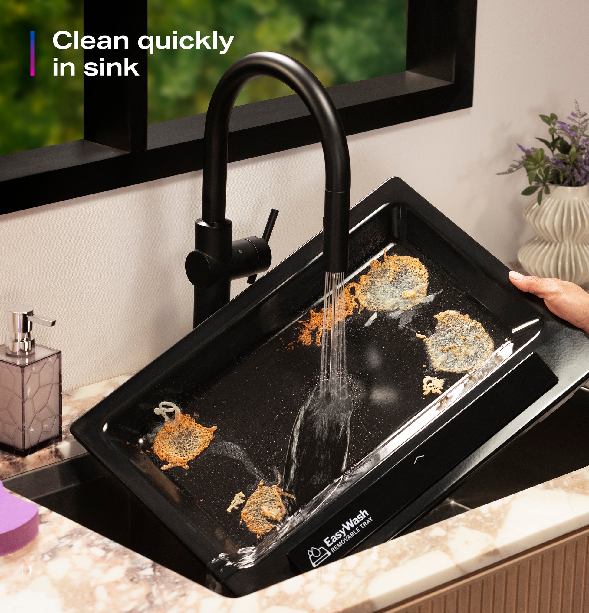 Person rinsing a dirty black EasyWash™ oven tray under a modern black kitchen faucet in sink area.