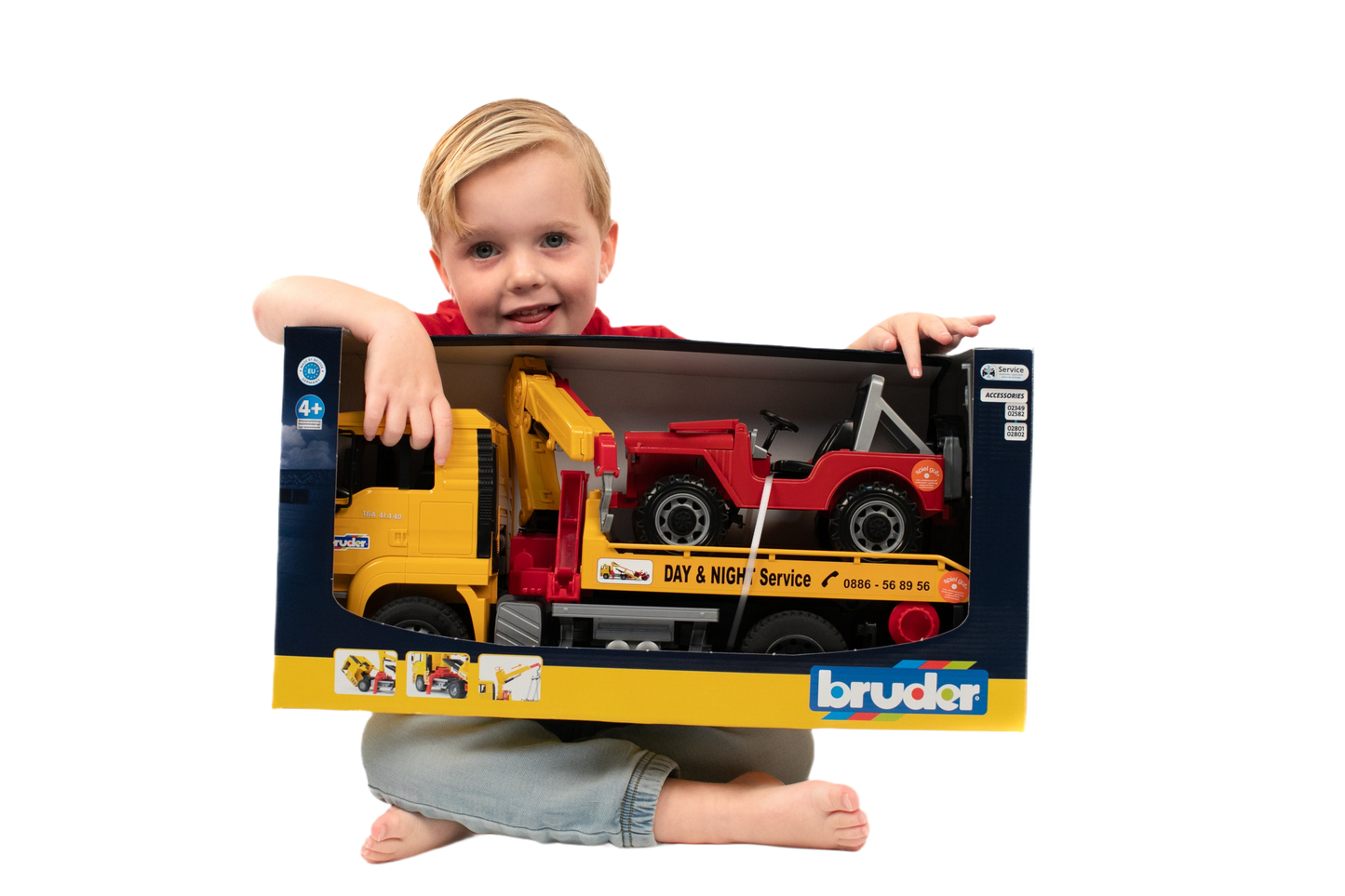 Child holding Bruder toy MAN TGA yellow tow truck carrying red cross country vehicle in display packaging