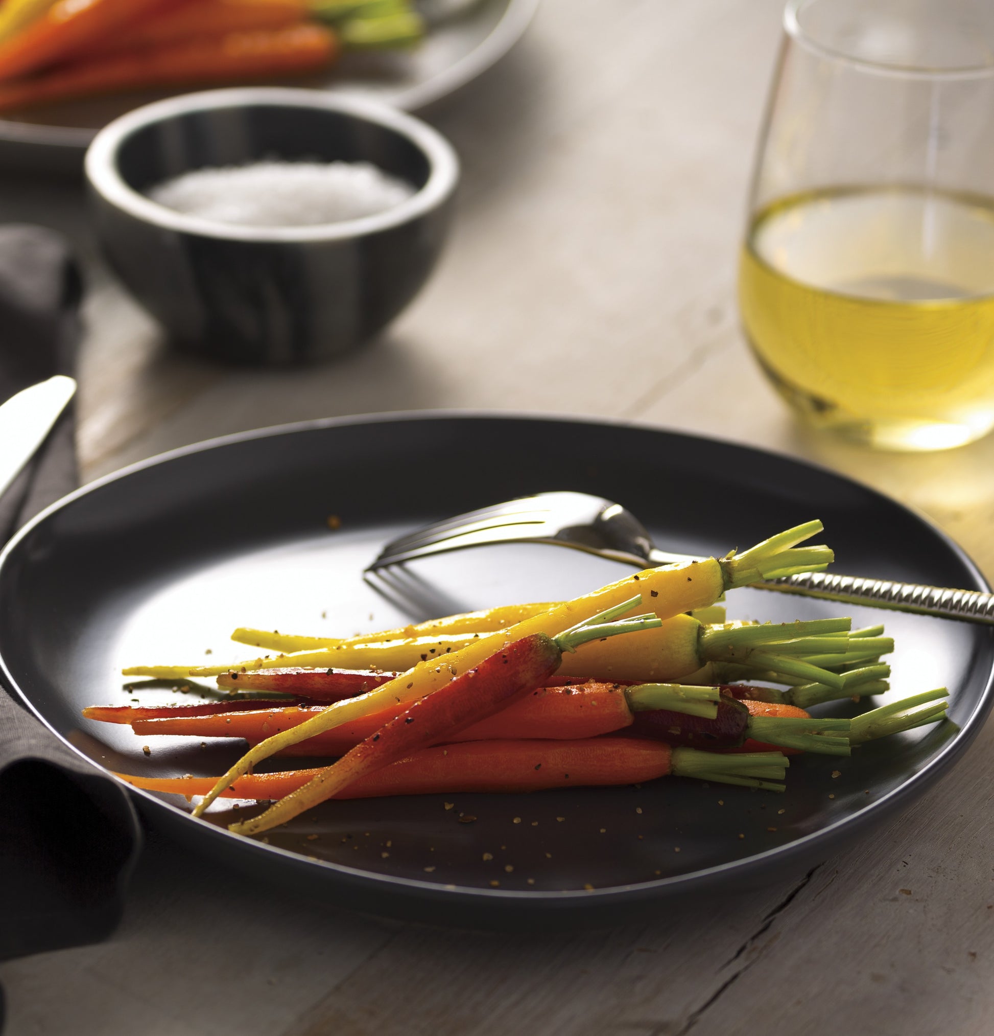 Black plate with seasoned cooked rainbow carrots beside a glass of white wine and small bowl of salt