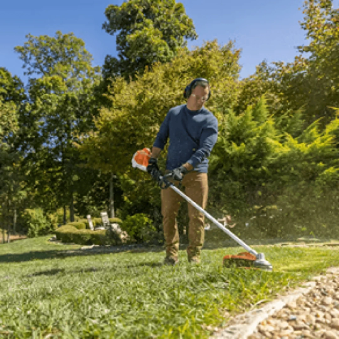 Man wearing protective gear using a straight shaft trimmer for outdoor lawn care along a gravel edge