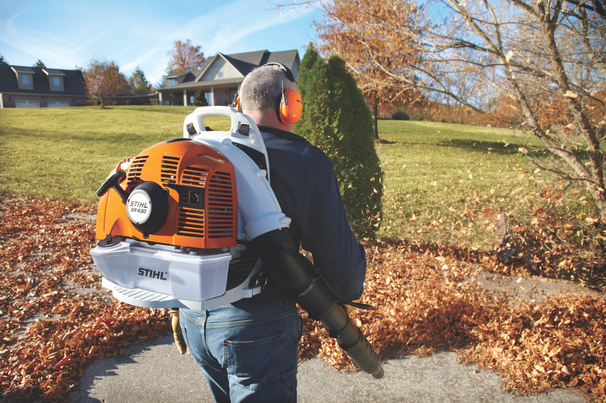 Man wearing ear protection using orange and white STIHL backpack blower to clear fallen leaves on paved path
