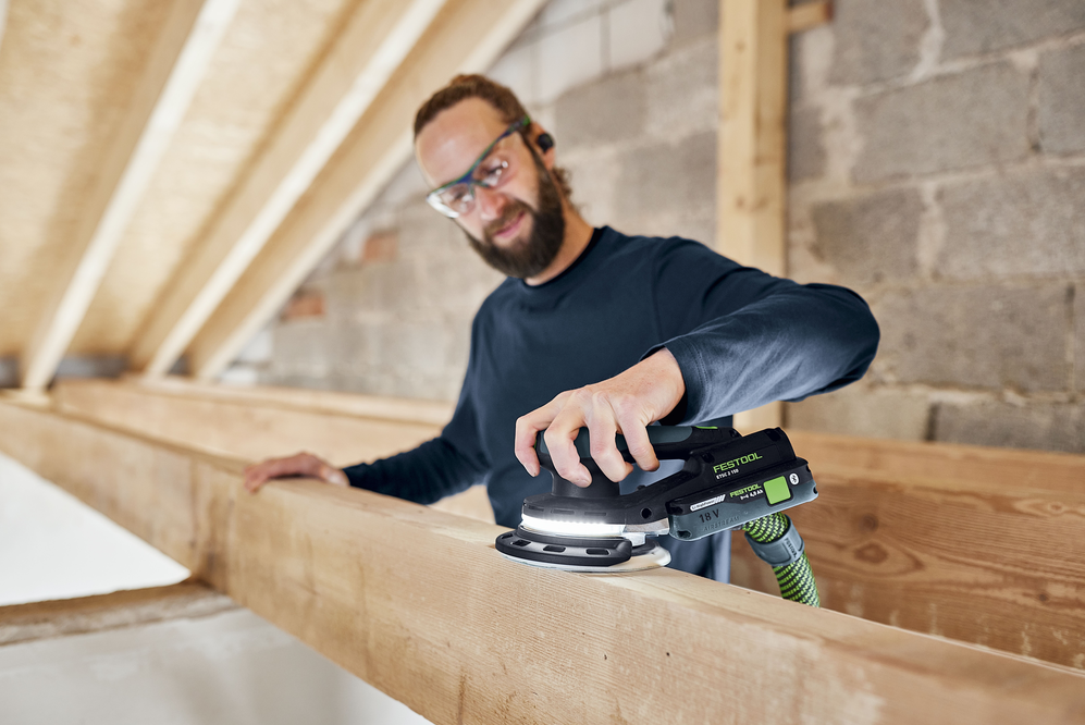 Man using cordless Festool orbital sander on wooden beam in a woodworking power tools and home improvement setting