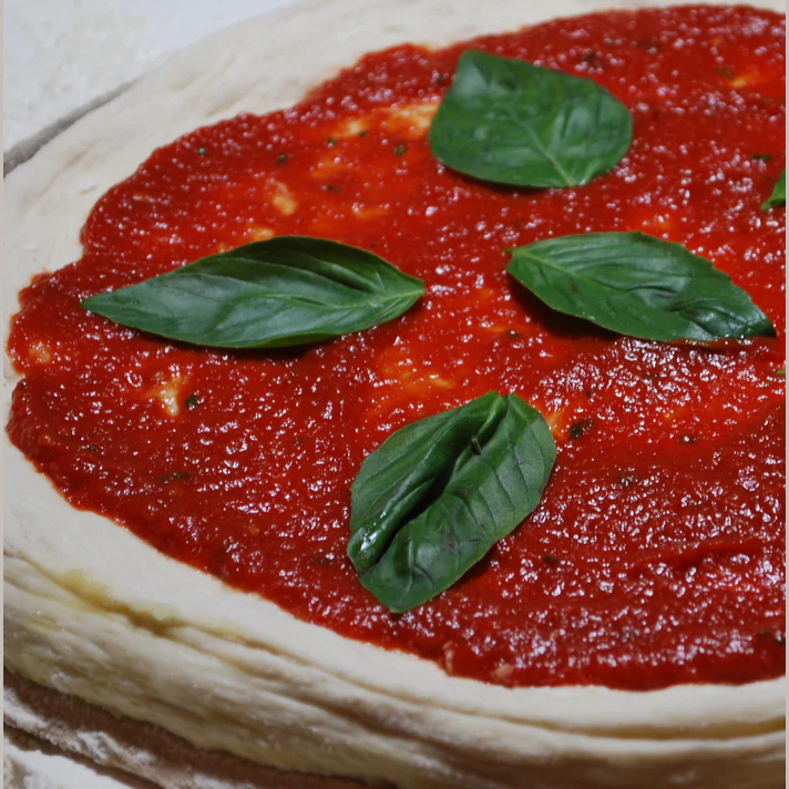 Close-up of fresh pizza dough spread with rich red sauce and topped with several green basil leaves in food and beverage collection