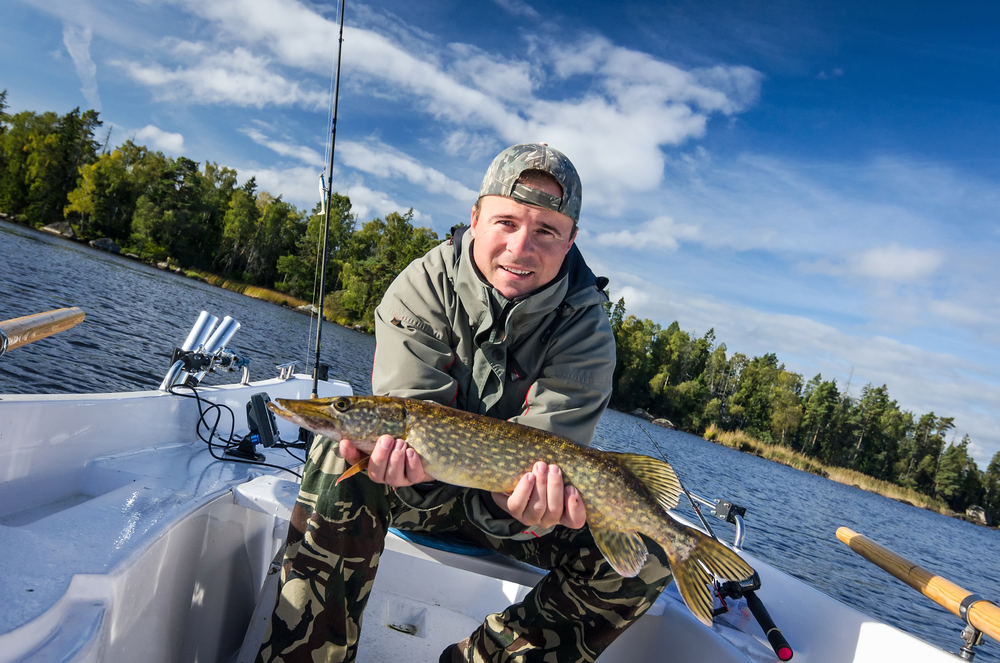Man wearing camouflage pants and a jacket sitting in a small boat on a lake holding a large fish with trees and a partly cloudy sky in the background.