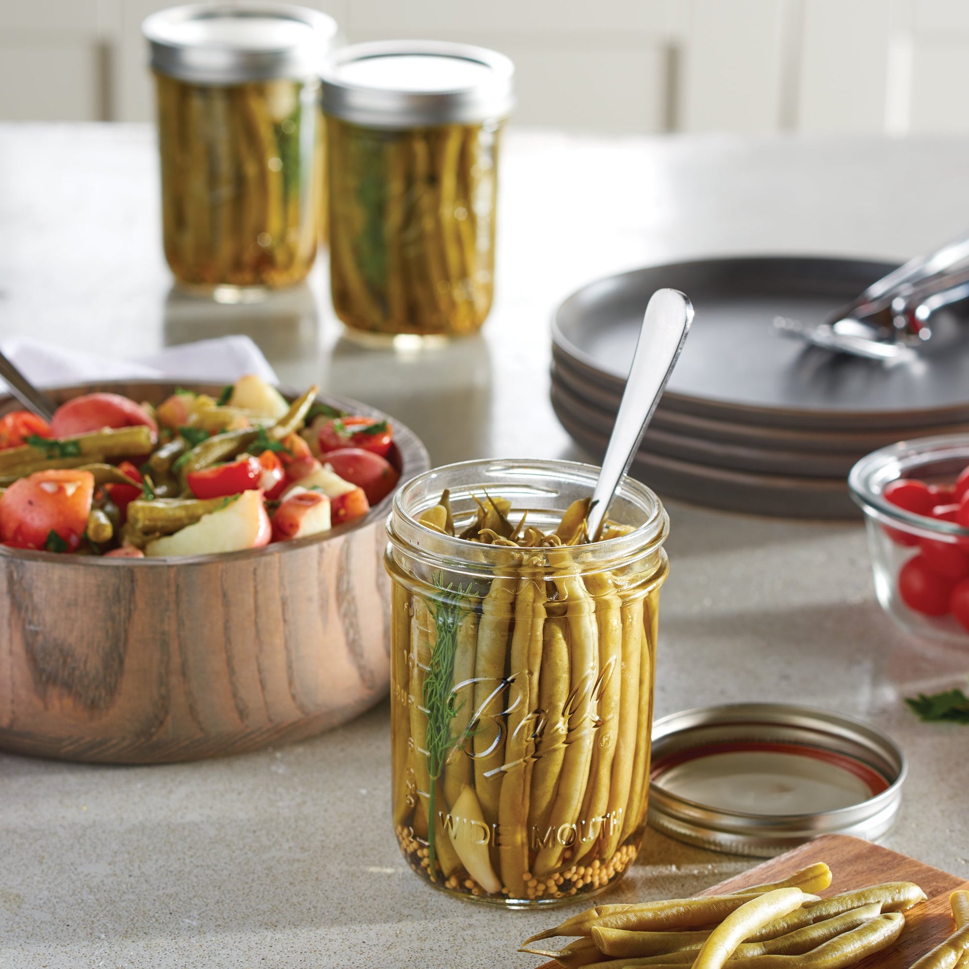 Clear glass wide mouth mason jar filled with pickled green beans and spoon, with salad bowl and plates in background