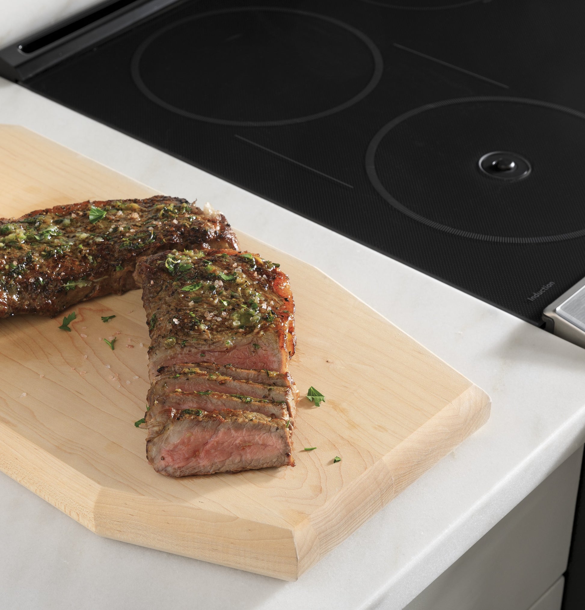 Sliced seasoned steak on wooden cutting board near black glass induction cooktop in kitchen and cookware setting