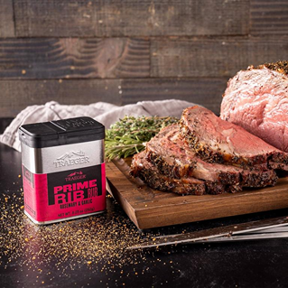 Square metal container of prime rib rub with rosemary and garlic seasoning next to sliced cooked prime rib on wooden board