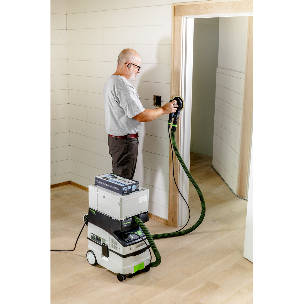 Man sanding a wood door frame using a Festool power sander and dust extractor in a home improvement setting