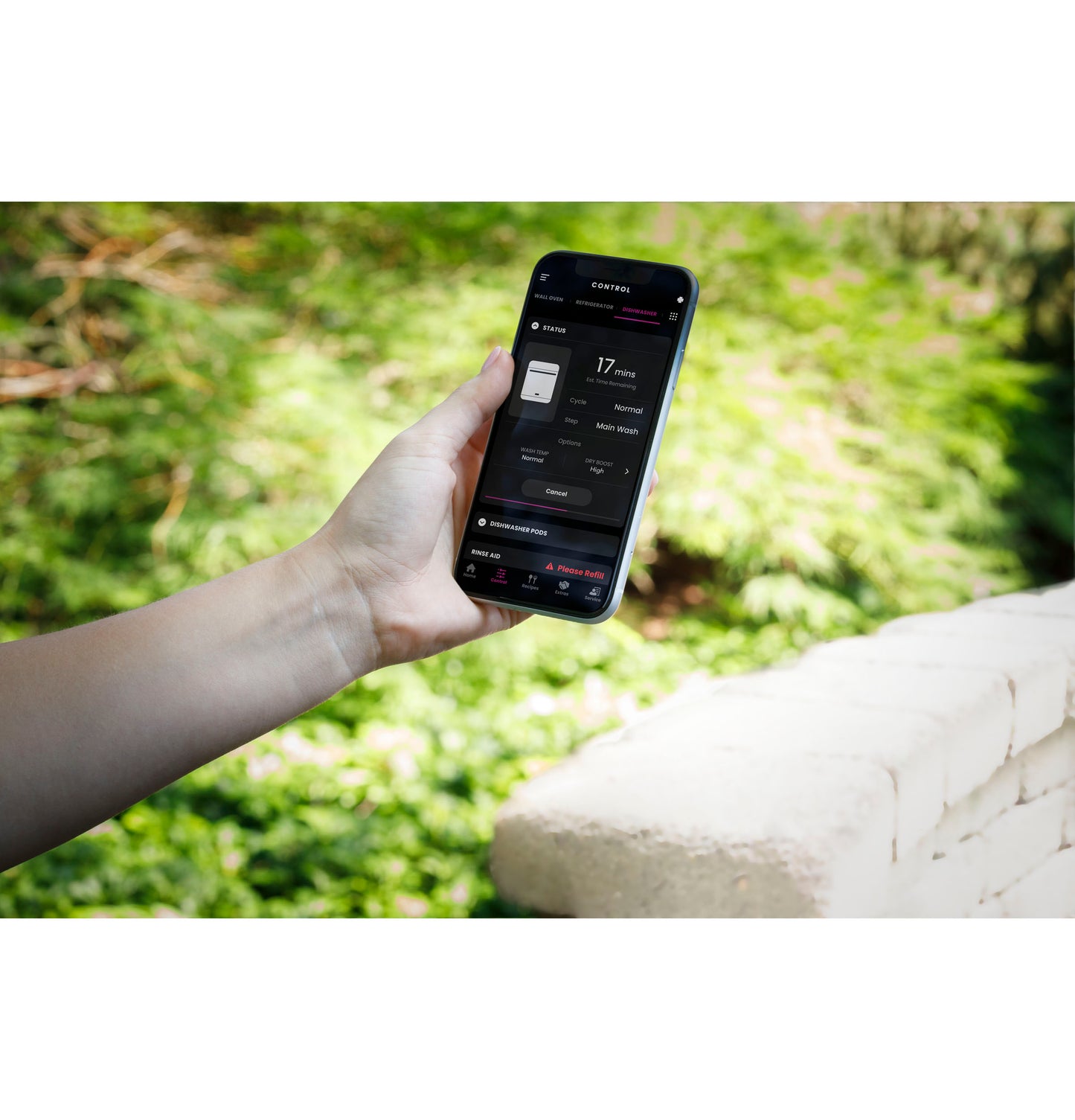 Hand holding smartphone displaying dishwasher control app outdoors with greenery in background in appliances collection