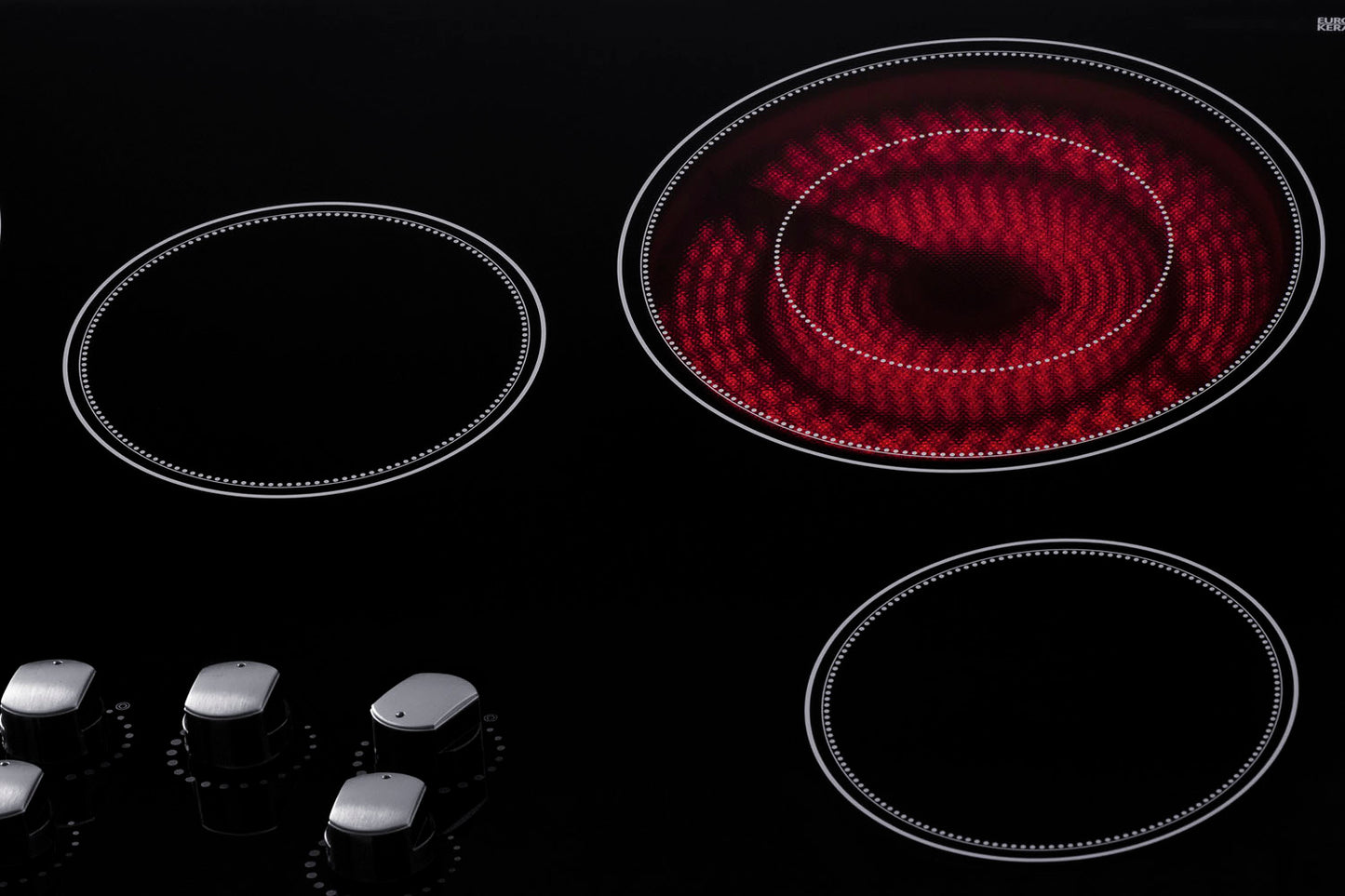 Black radiant cooktop with five burners, one active heating element glowing red, and four silver control knobs