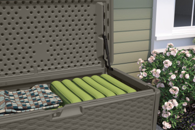 Outdoor resin wicker deck box with hinged lid storing green pool floats and folded towel by house siding and flowers