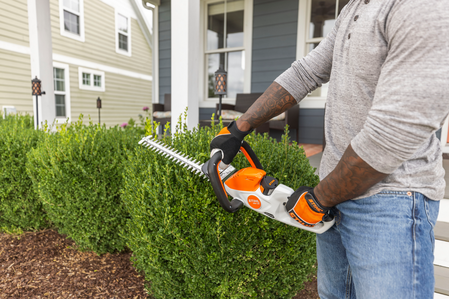 Person using orange and white electric hedge trimmer to shape green bushes in residential garden setting