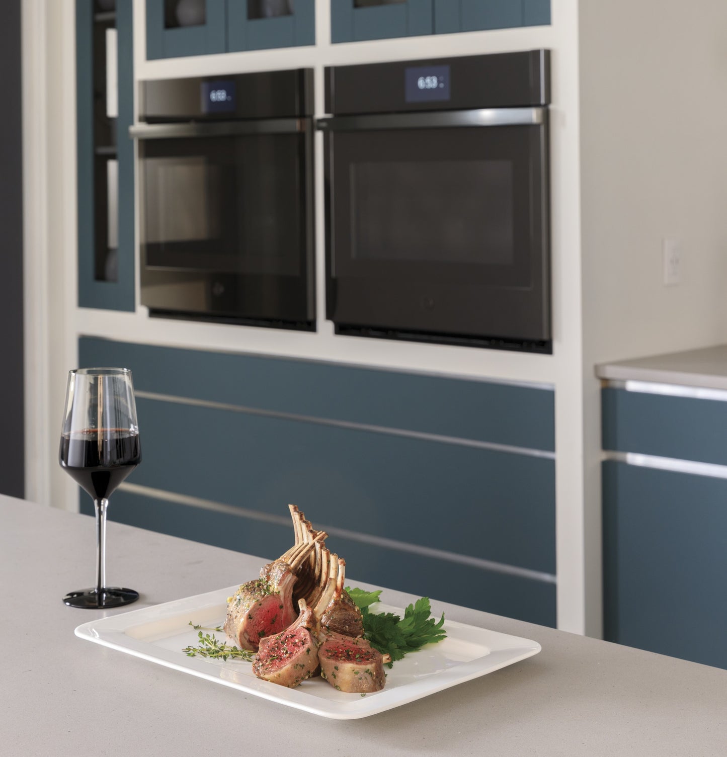 Plate of cooked lamb chops and herbs on kitchen island with double built-in wall ovens and wine glass nearby