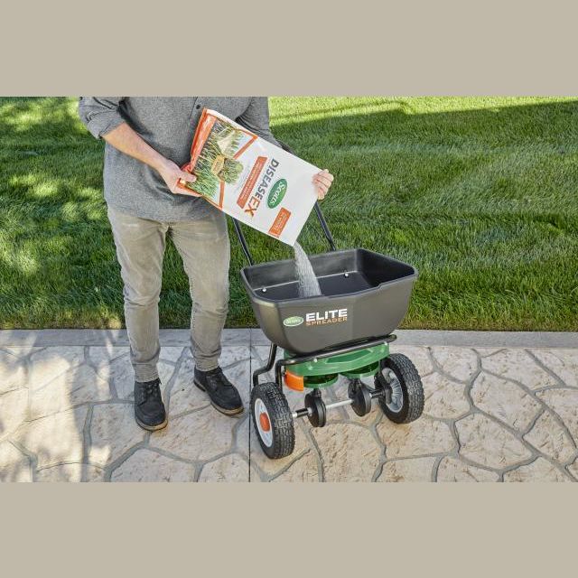Person pouring Scotts DiseaseEX lawn fungicide into a black and green broadcast spreader on patio in lawn and garden setting