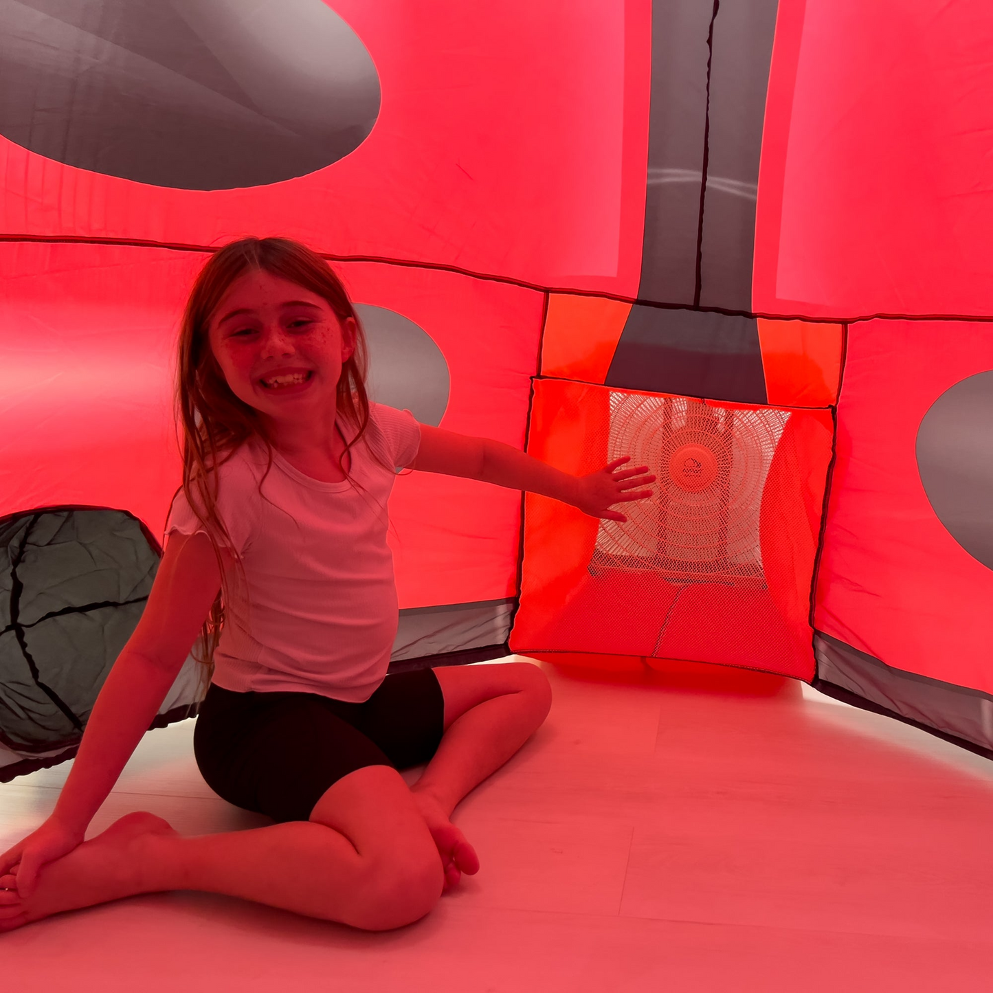 Smiling girl sitting inside a red and black inflatable ladybug play tent with mesh window for ventilation