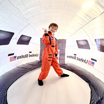 Child dressed in orange astronaut suit playing inside a white inflatable space shuttle tent with US flag decals
