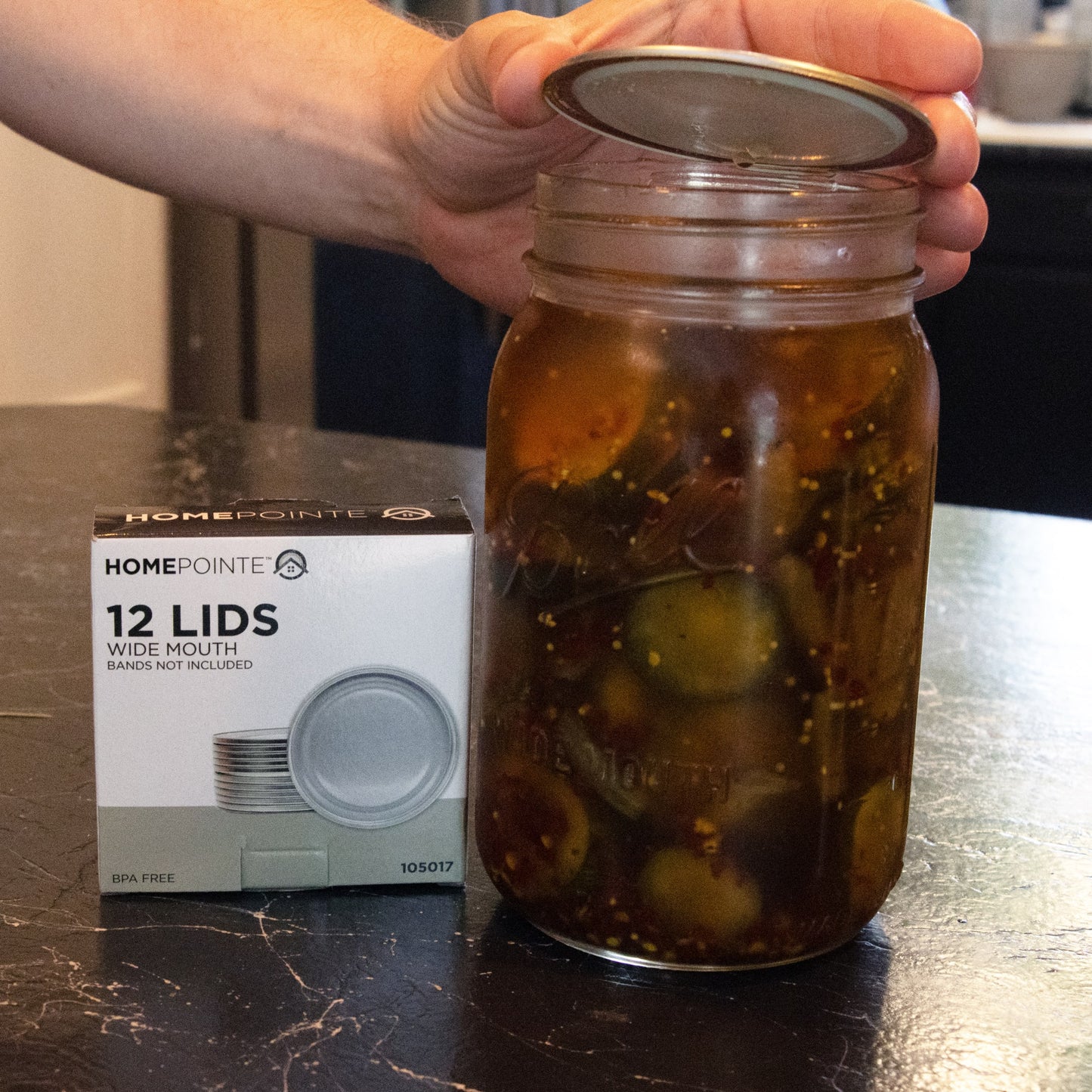 Hand placing a wide-mouth lid on a glass jar of pickles next to a box of 12 lids in kitchenware collection