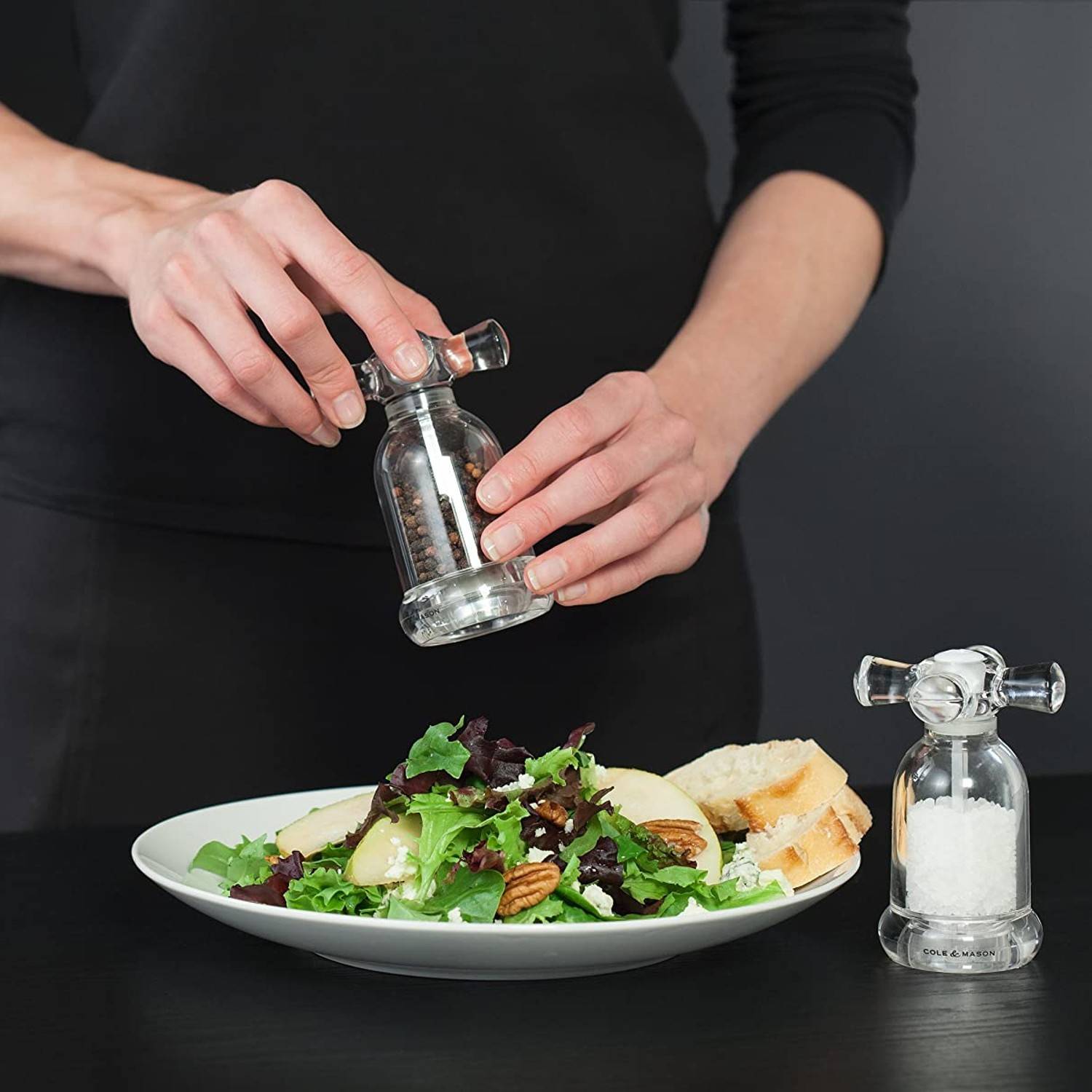 Person grinding pepper from clear acrylic mill with vintage-style handle over fresh salad in kitchenware setting