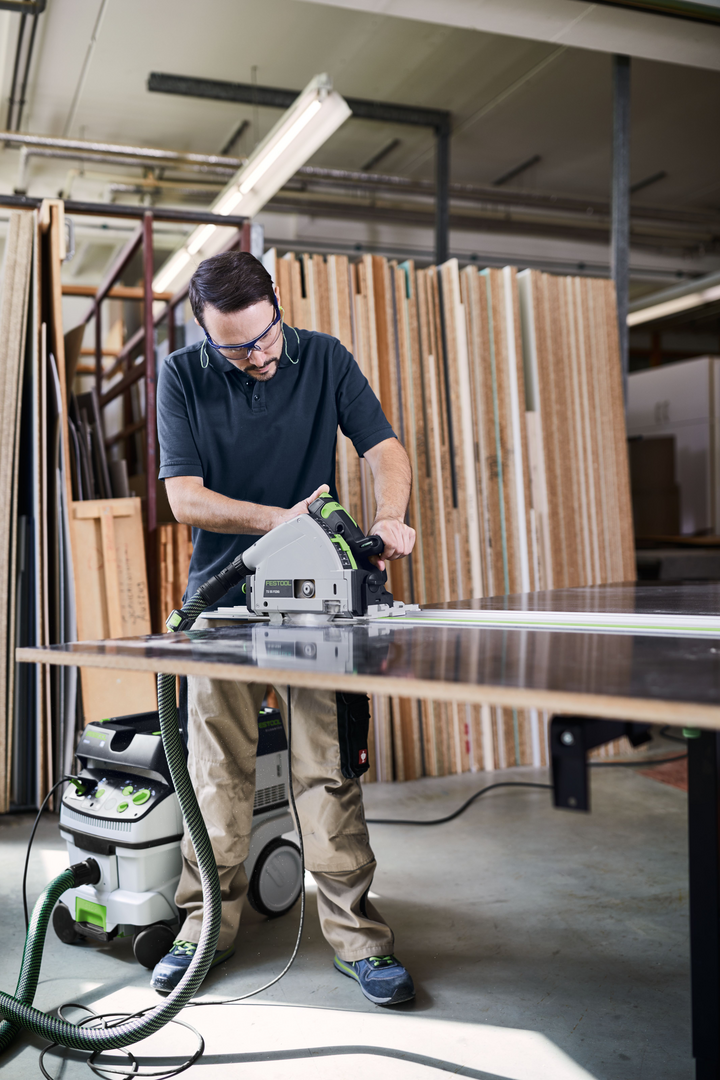 Man in safety glasses using Festool power tool connected to HEPA dust extractor in woodworking workshop