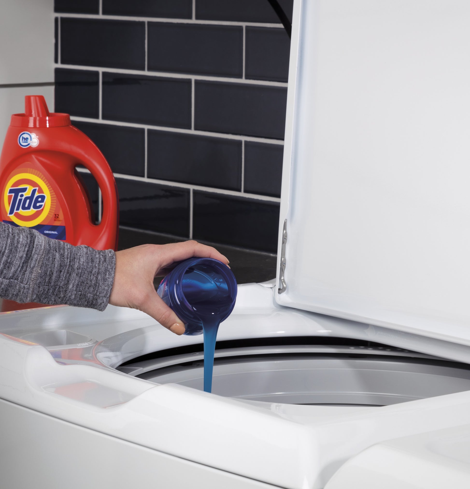 Person pouring blue liquid detergent into white top load washer with open lid in modern laundry space
