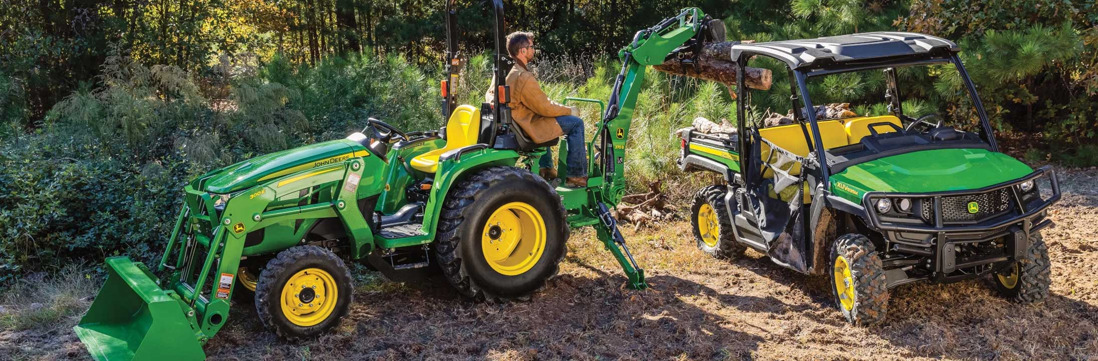 Person operating a green and yellow tractor with a front loader and backhoe attachment next to a green and yellow utility vehicle on a dirt path surrounded by trees.