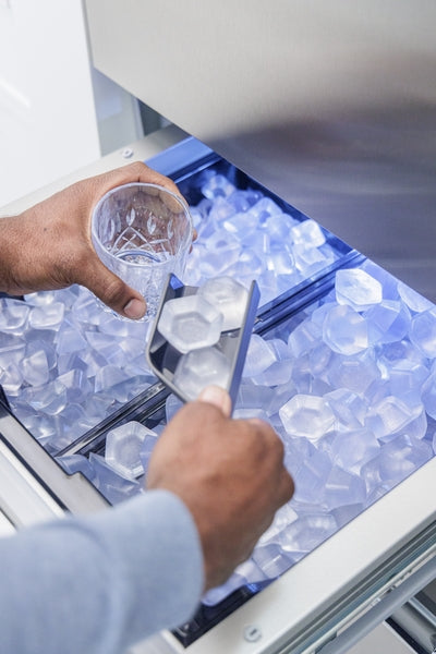 Close-up of person scooping large hexagonal ice cubes into glass from bottom freezer ice bin of stainless steel refrigerator