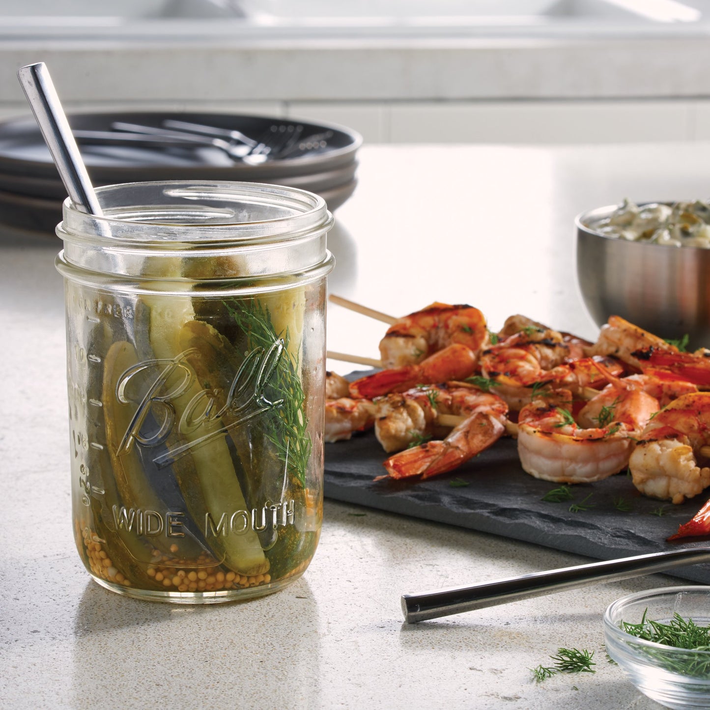 Clear glass wide mouth mason jar filled with pickles and herbs next to grilled shrimp skewers on kitchen counter