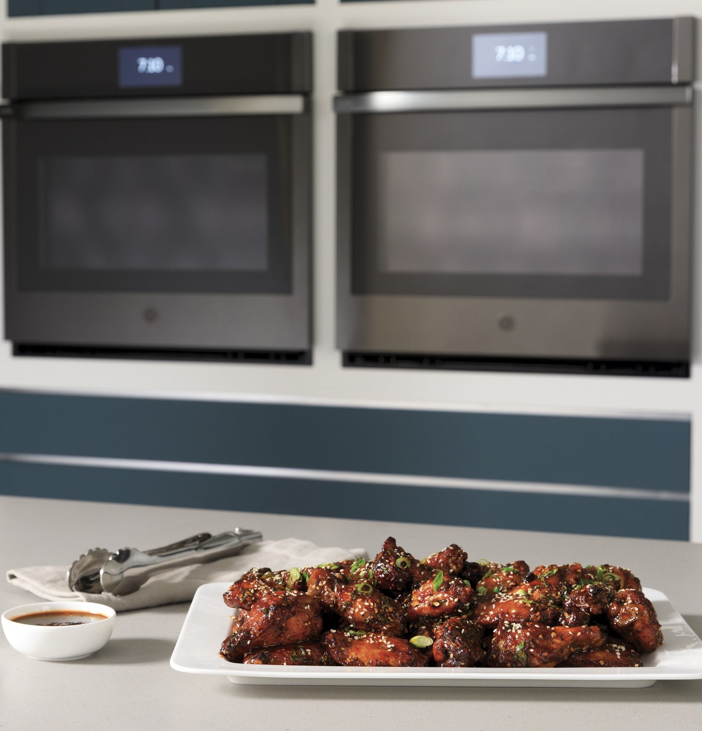 Plate of glazed chicken wings garnished with green onions on kitchen island beneath built-in convection double wall ovens