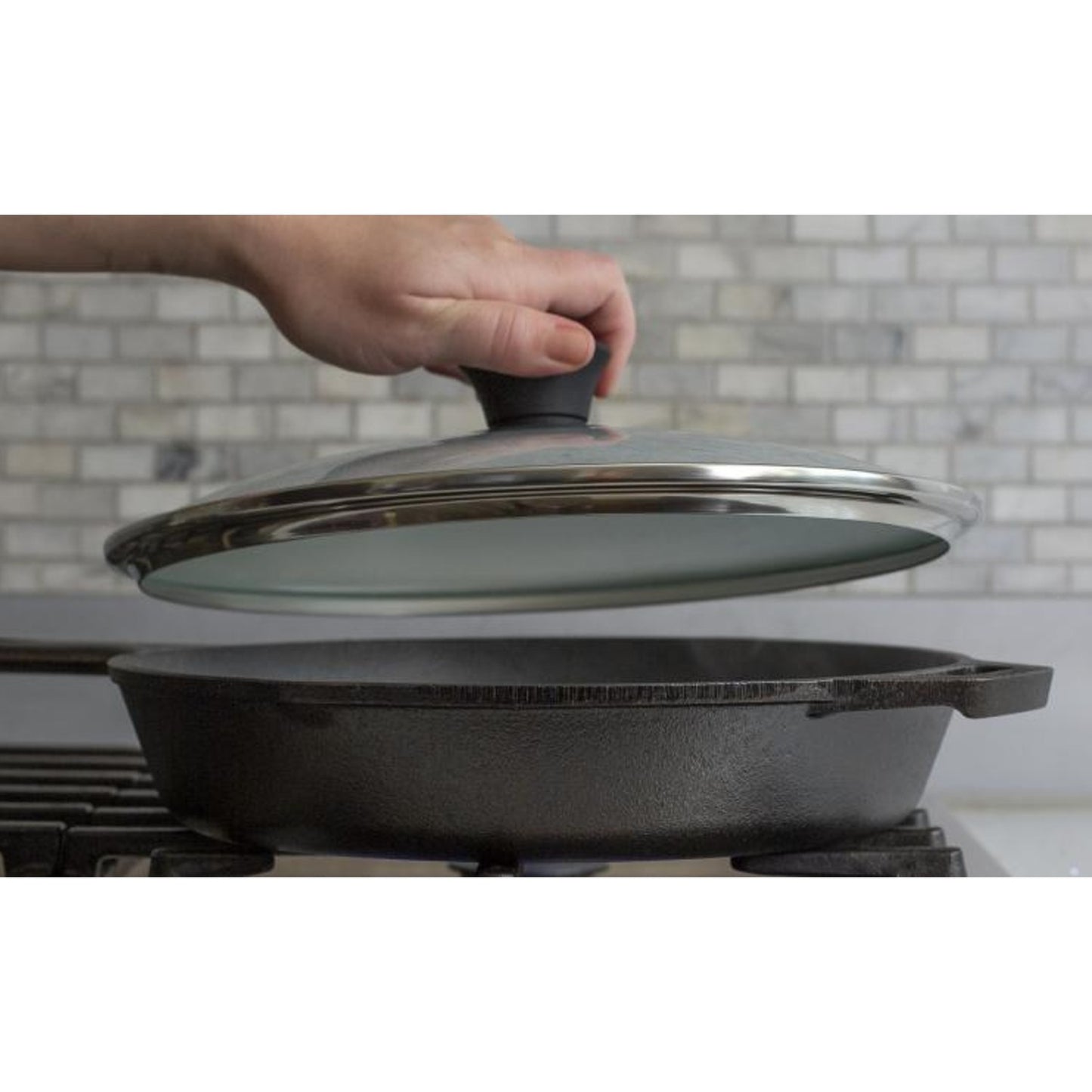 Hand lifting round tempered glass lid with black knob and stainless steel rim from cast iron skillet in kitchenware collection