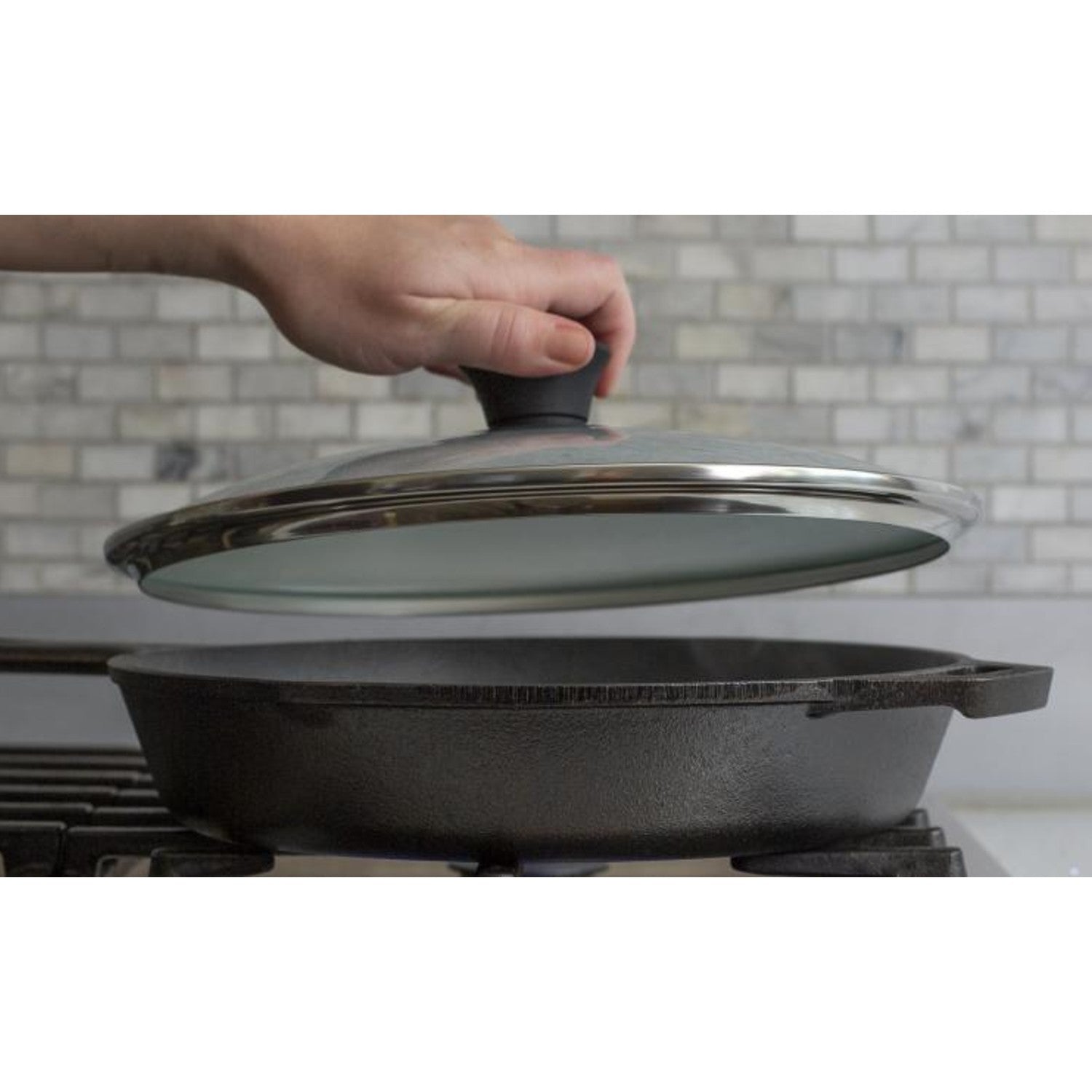 Hand lifting round tempered glass lid with black knob and stainless steel rim from cast iron skillet in kitchenware collection