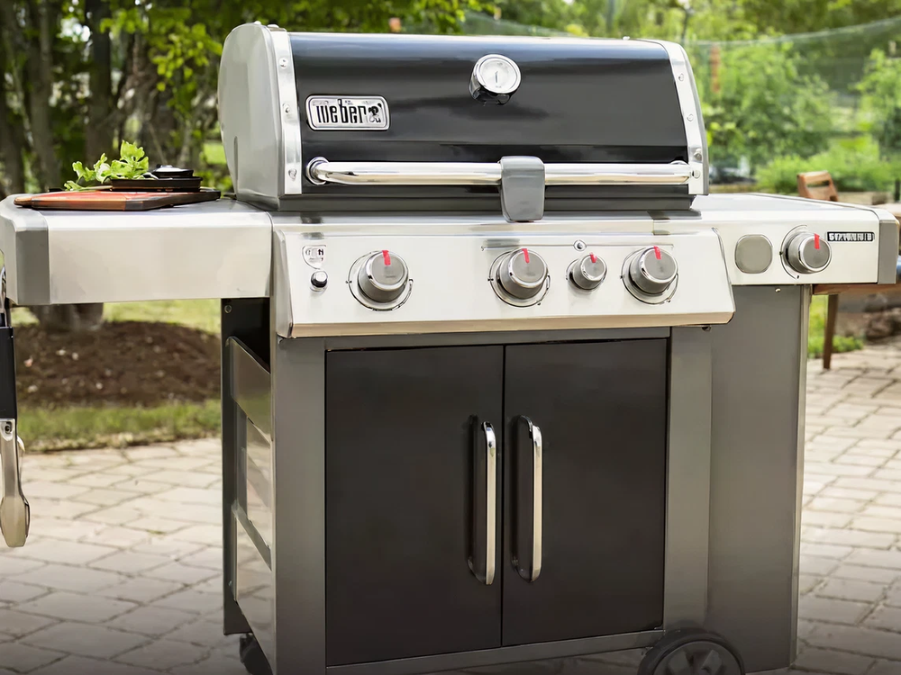 Outdoor stainless steel grill with four control knobs and double doors on a stone patio surrounded by greenery.